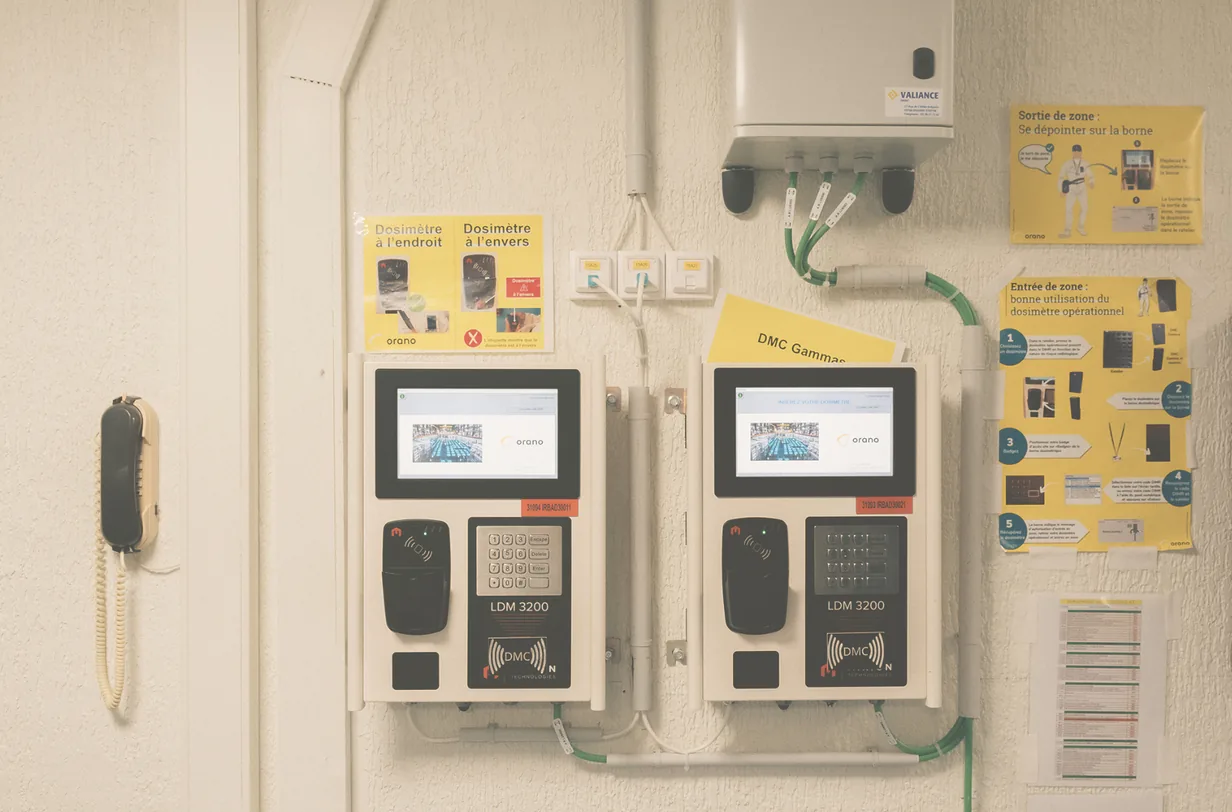 Zwei Dosimeter-Terminals mit Bildschirmen und gelben Hinweistafeln an einer Wand in La Hague.